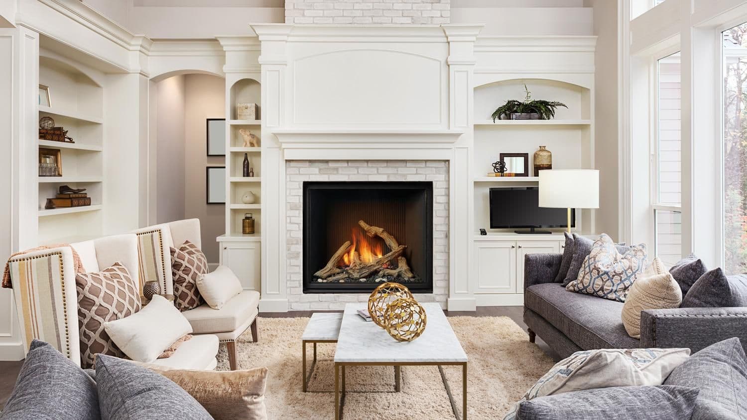 Elegant living room with a cozy lit fireplace centered between built-in shelves. Neutral-tone sofas and plush pillows create a welcoming, sophisticated ambiance.