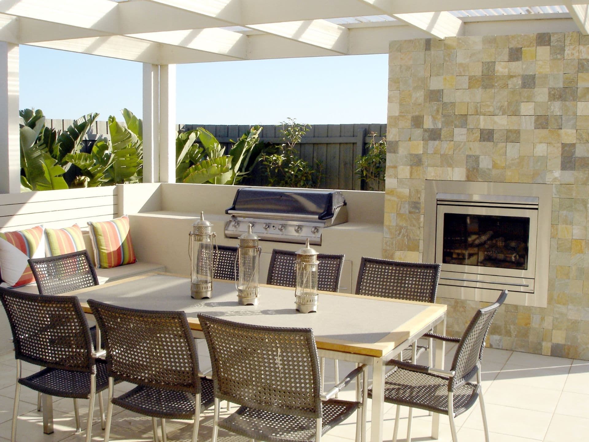 Outdoor patio with a tiled wall and fireplace, a dining table with wicker chairs, a built-in grill, colorful cushions, and lush green plants.