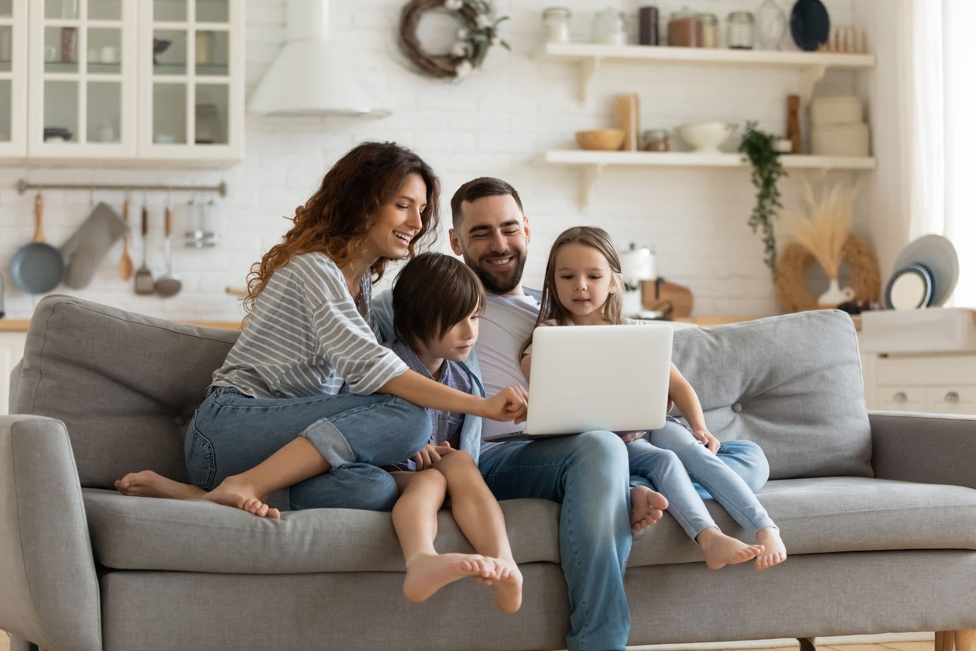 A smiling family of four sits on a cozy gray sofa, gathered around a laptop. The living room backdrop is a bright, modern kitchen, creating a warm atmosphere.