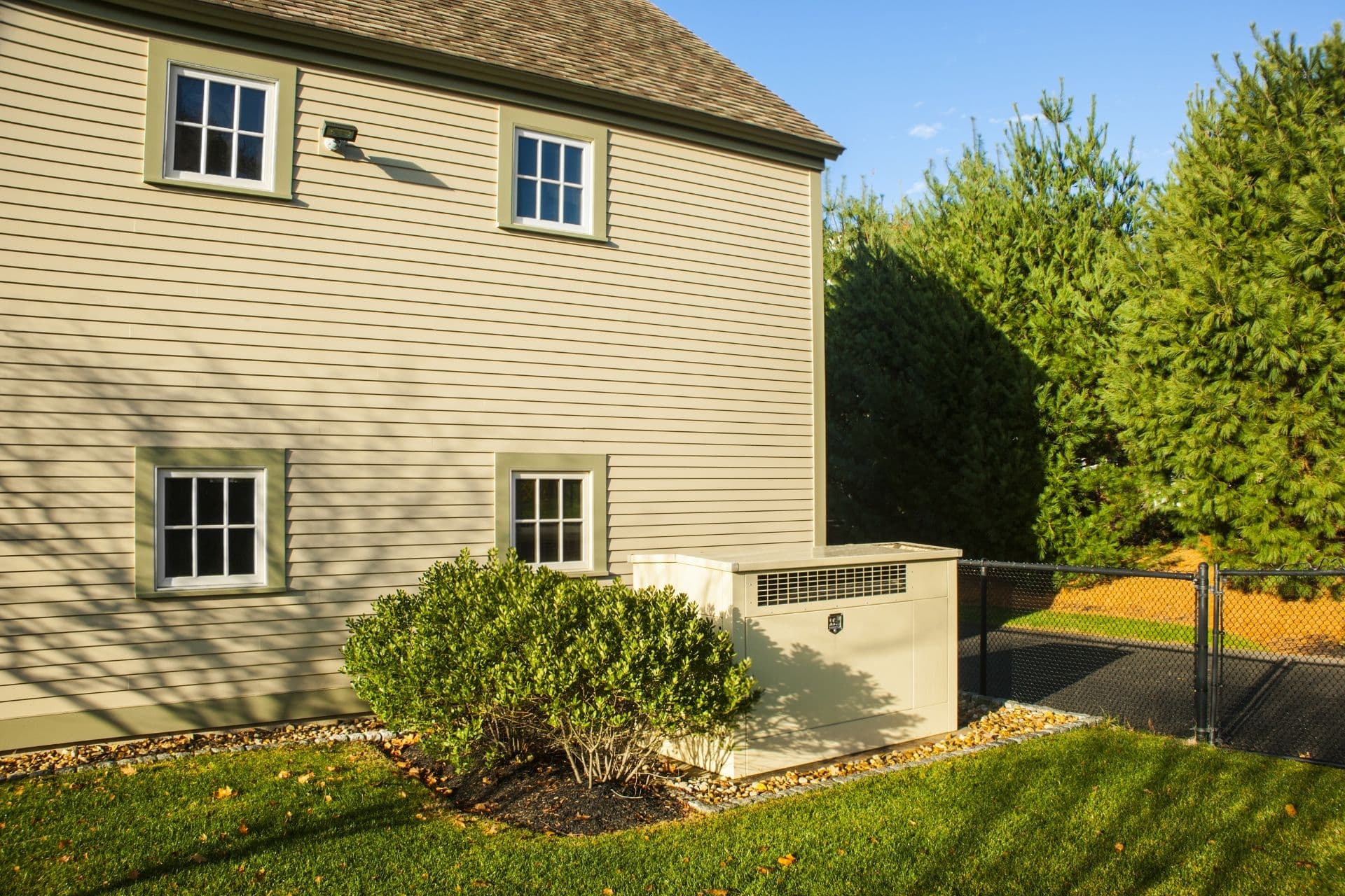 A beige house exterior with three windows, surrounded by green shrubs and grass. A large light-colored generator is next to a black chain-link fence.