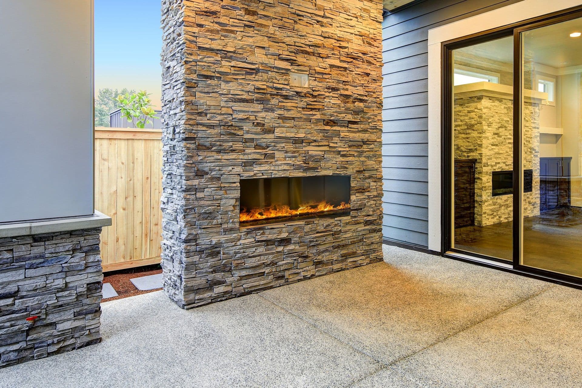 Modern outdoor patio with a stone-clad wall and built-in linear fireplace. Large sliding glass doors reveal a cozy interior room. Warm, inviting ambiance.