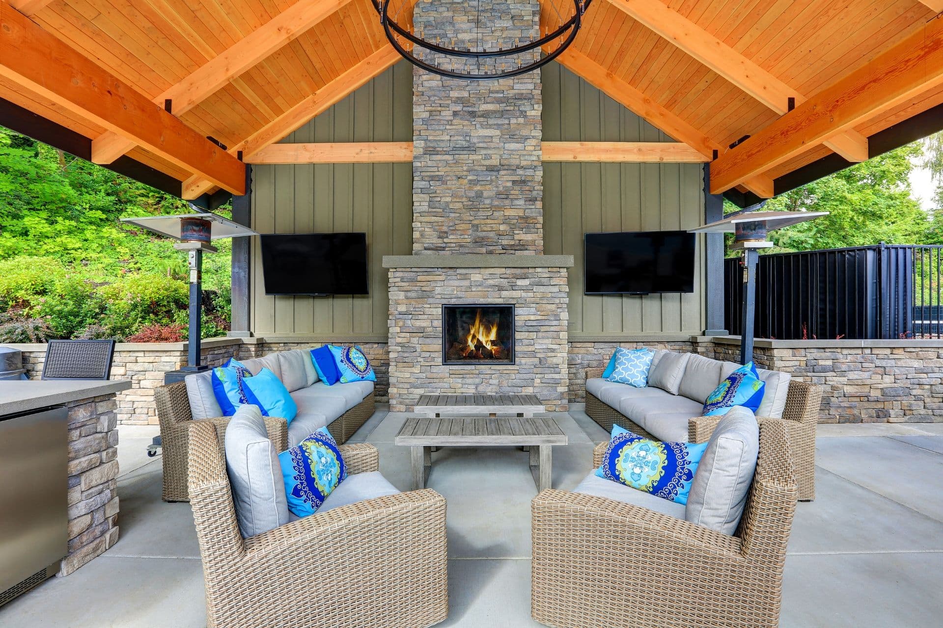 Outdoor patio with wooden ceiling, stone fireplace, and wicker furniture. Cushions in blue tones create a cozy, inviting atmosphere. Two TVs are mounted on the walls.