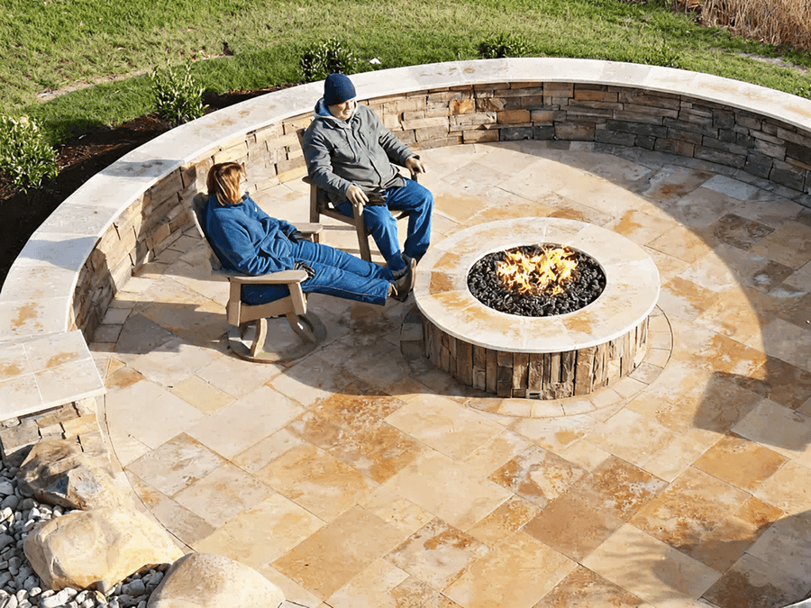 Two people sit on a stone patio with a circular fire pit in the center. The scene is tranquil and surrounded by green grass and small plants.