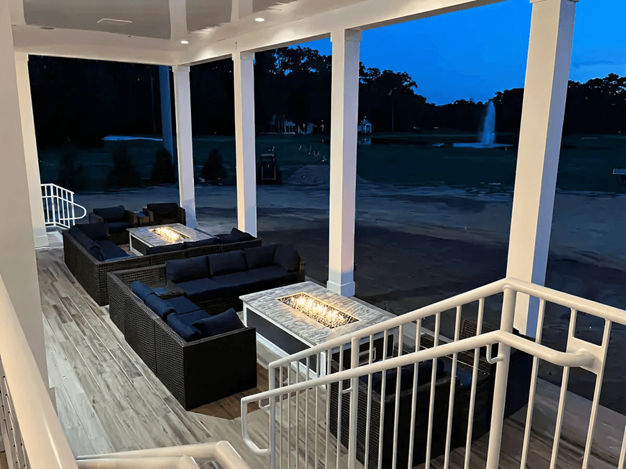 Outdoor patio at dusk with cozy wicker sofas, fire pits on wooden flooring, white railing, and a view of a golf course and fountain in the distance.