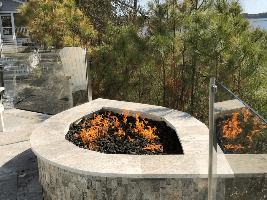 A stone fire pit with glowing orange flames set on a patio, surrounded by glass panels and overlooking a lush forest and lake backdrop.
