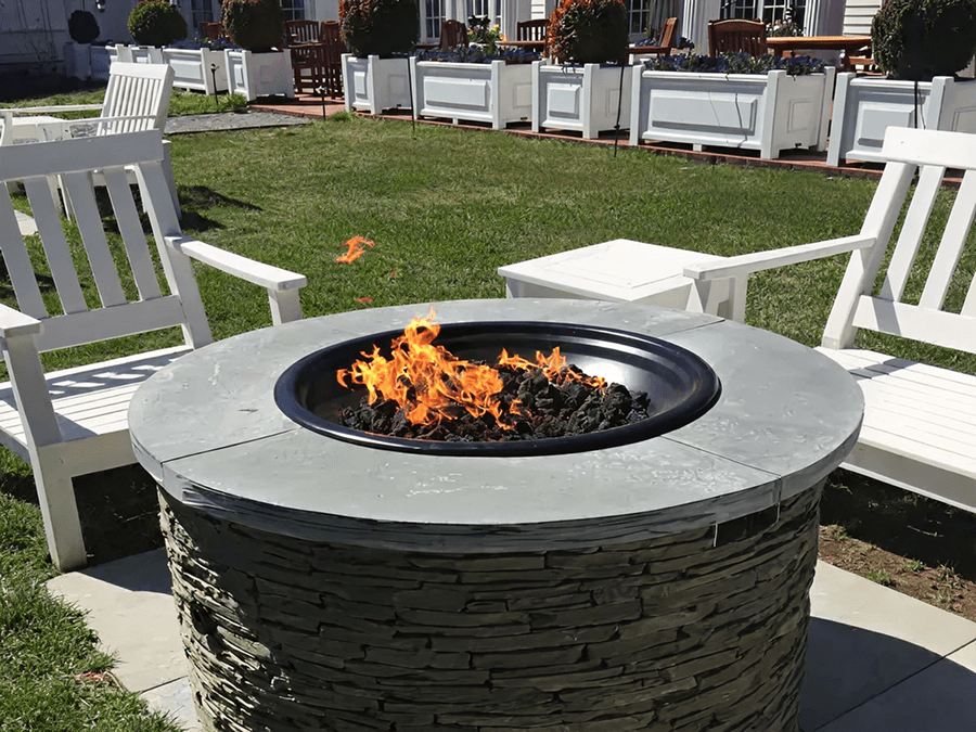 A round stone fire pit with bright orange flames is situated on grass. White wooden benches encircle it, creating a cozy outdoor seating area.