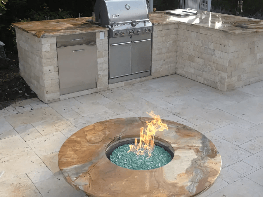 Outdoor kitchen with a stone counter, stainless steel grill, and a circular fire pit with glass stones, set on a tiled patio.