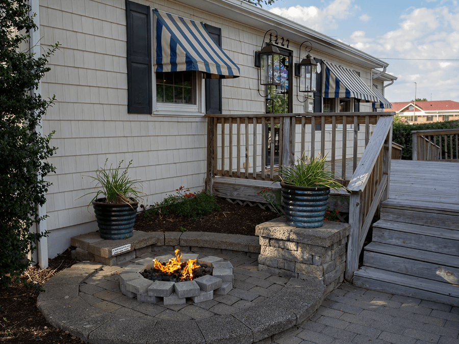 A cozy outdoor patio features a small stone fire pit surrounded by paving stones. Blue-striped awnings and potted plants create a relaxed atmosphere.