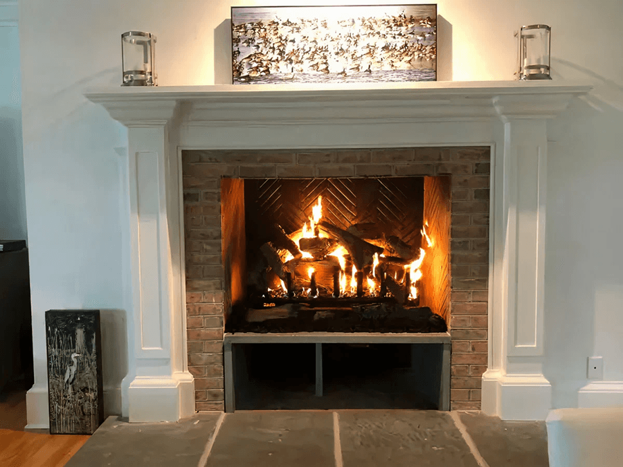 A cozy lit fireplace framed by white mantel and brick interior. A decorative picture and lanterns sit on the mantel, exuding warmth and comfort.