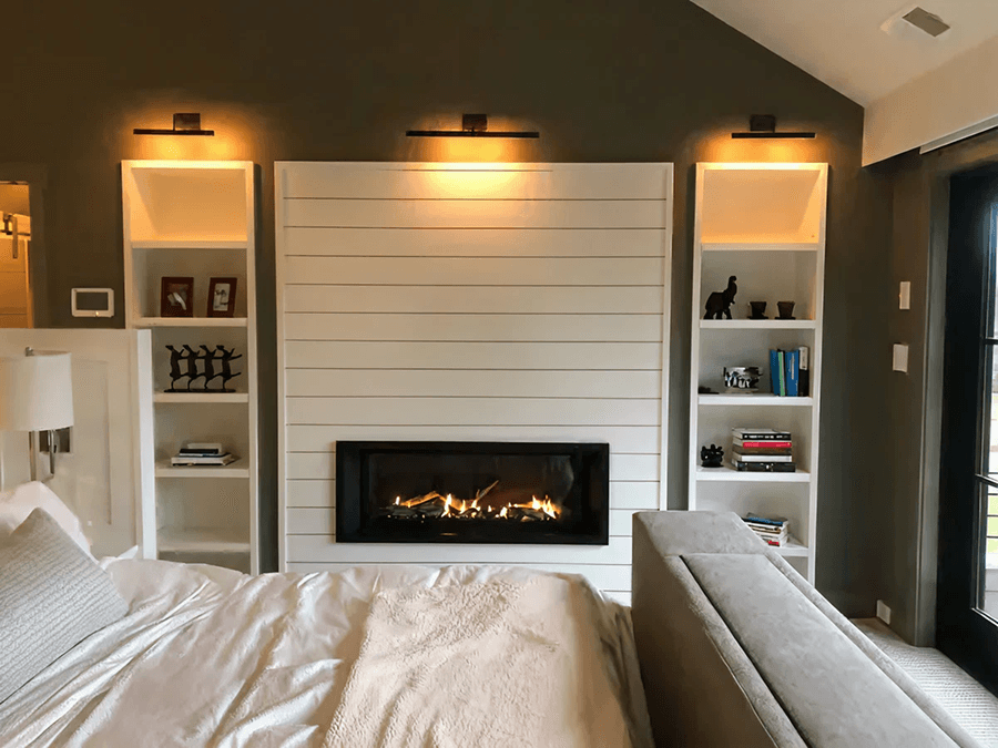 Cozy bedroom with a modern fireplace set in a white shiplap wall. Shelves on either side hold books and decor. Warm lighting creates a serene atmosphere.