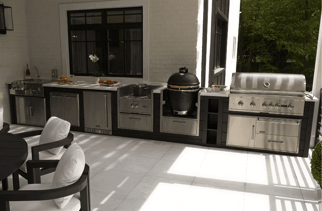 Modern outdoor kitchen with stainless steel appliances, including a grill, smoker, and oven. White brick walls and sleek patio furniture create an elegant ambiance.
