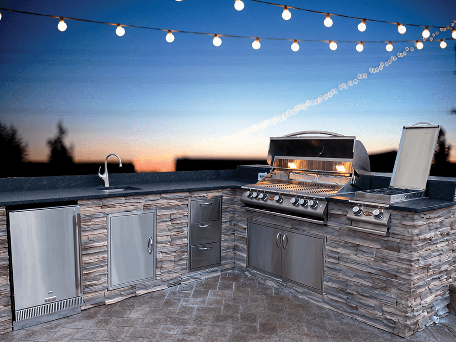 Outdoor kitchen with stainless steel appliances, including a grill and side burner, against a stone counter at dusk. String lights hang above, creating a cozy ambiance.