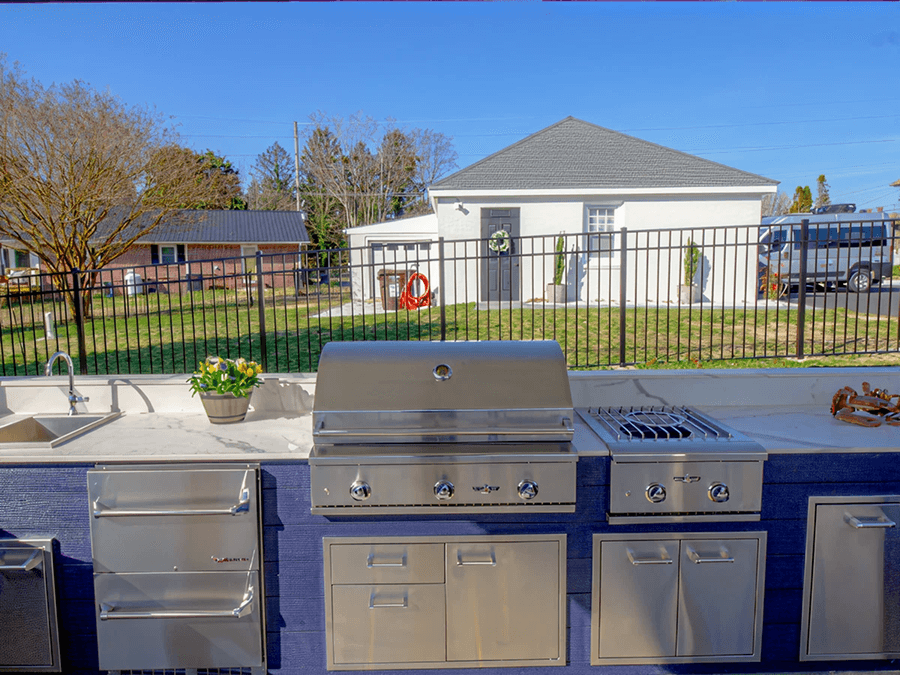 Outdoor kitchen with stainless steel grill, stove, and drawers against a blue counter. Potted plant and sink on left. White building and trees in background.