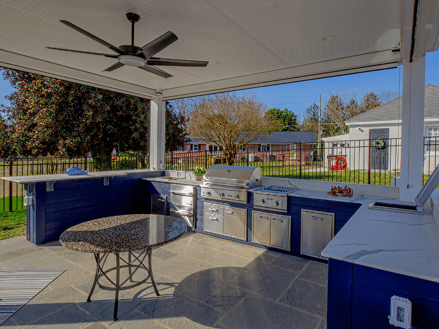 Outdoor kitchen with stainless steel appliances, including a grill, under a covered patio. A ceiling fan above and a round stone table in the foreground. Trees and houses are visible in the background.
