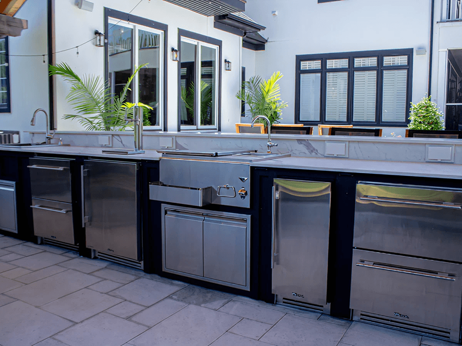 Outdoor kitchen with stainless steel appliances, including a sink and fridge, set against a modern home. Potted plants add greenery.