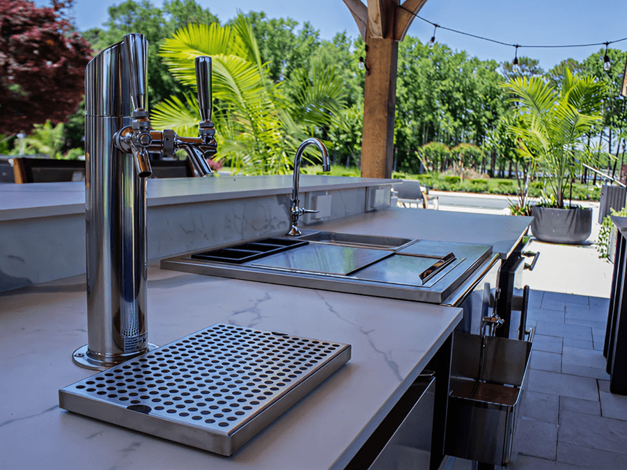 Outdoor kitchen with a sleek marble countertop, chrome beer tap, and built-in sink. Surrounded by potted plants under a bright blue sky.