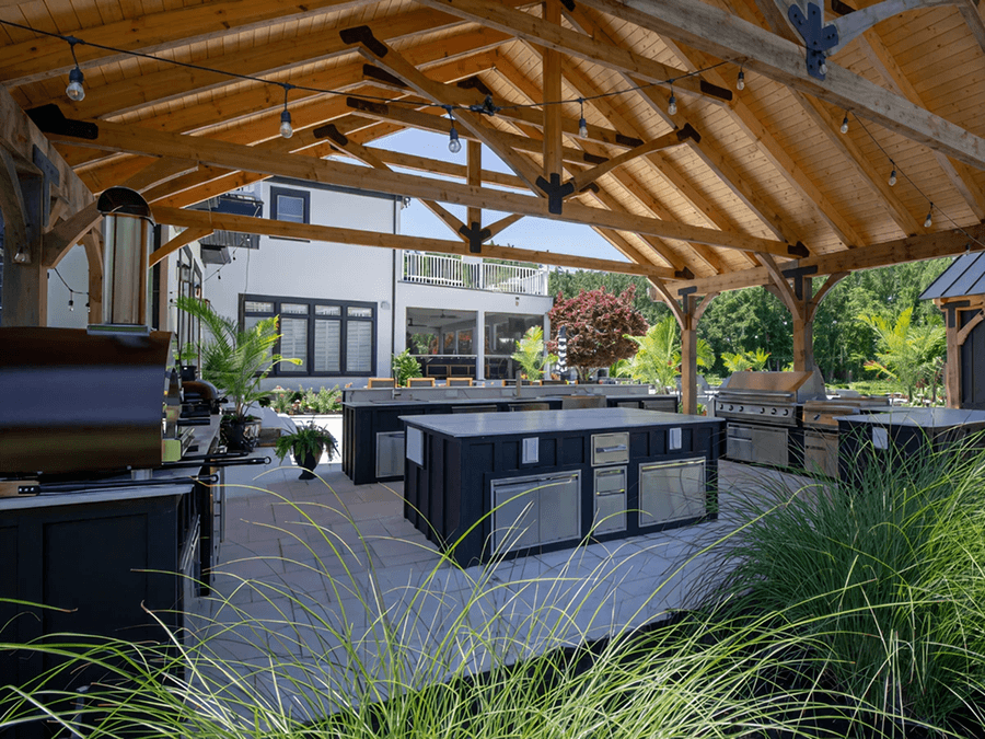 Expansive outdoor kitchen under a wooden pergola. Features modern appliances, lush greenery, and a view of a contemporary house.