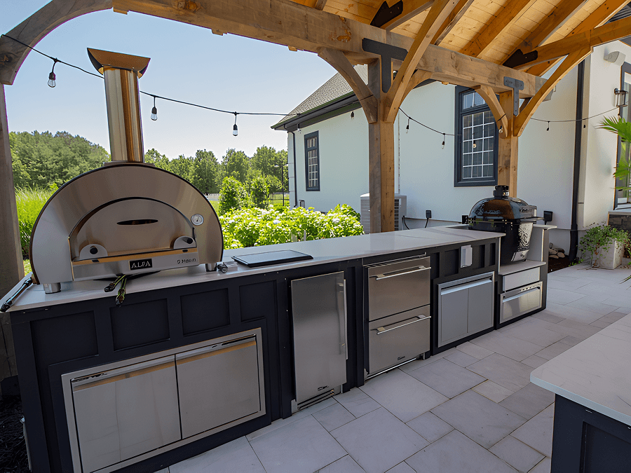 Outdoor kitchen with a pizza oven, grill, and stainless steel appliances under a wooden pergola, surrounded by greenery and a sunny atmosphere.