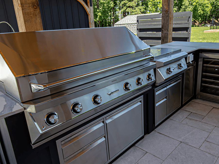 Sleek stainless steel outdoor kitchen with two large grills and storage drawers, set against a patio and green lawn.