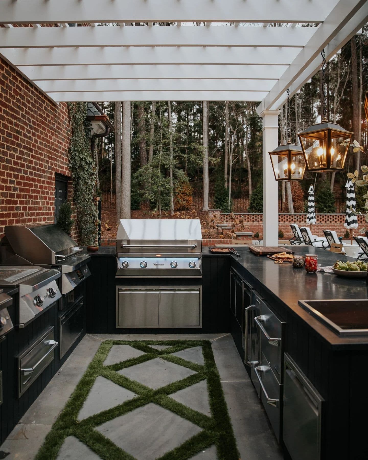 Outdoor kitchen with a modern grill under a white pergola. Surrounded by brick walls and tall trees, the area has a cozy and inviting atmosphere.