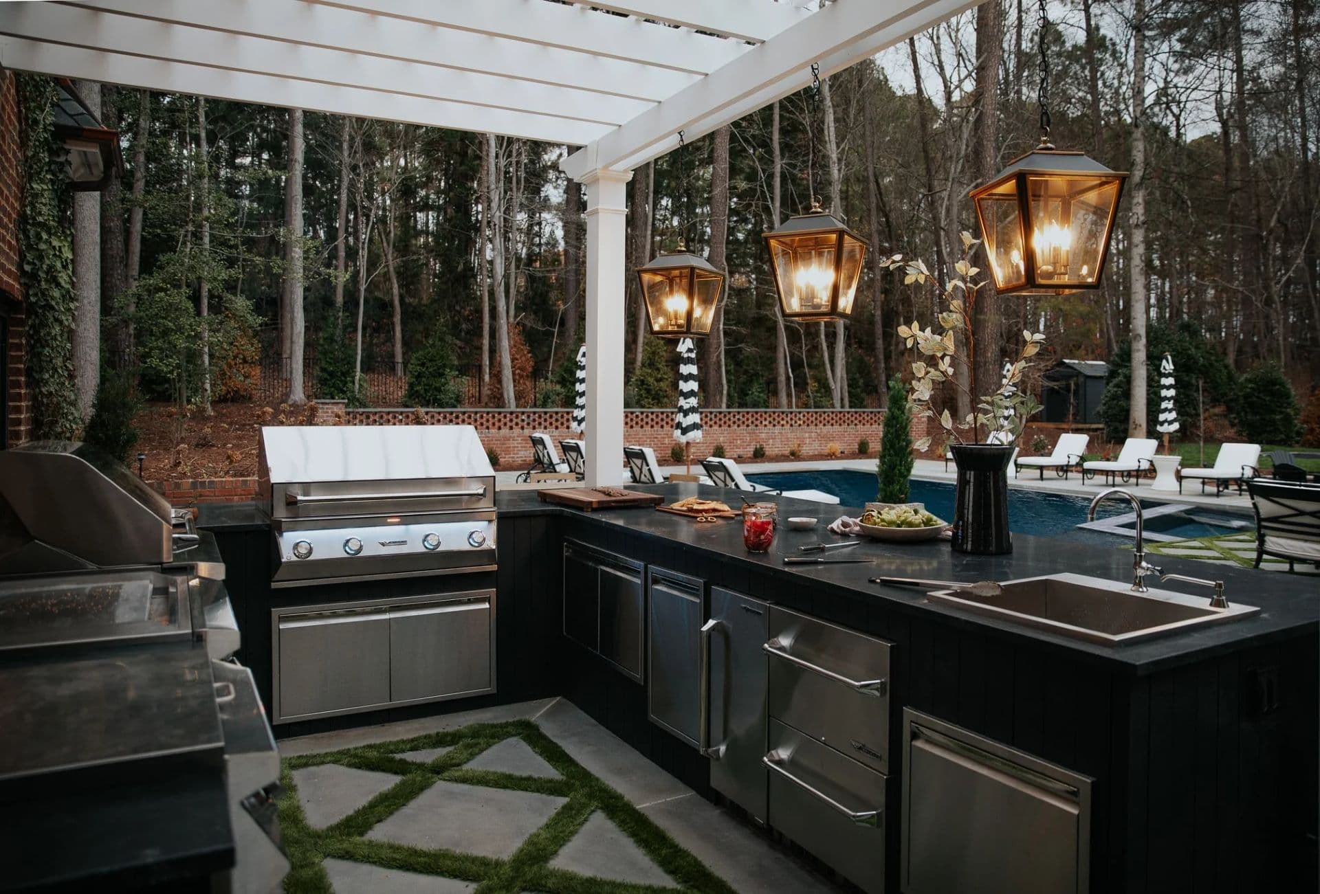 Outdoor kitchen with stainless steel appliances under a pergola, featuring warm lantern lights. View of a pool and forest, creating a cozy, inviting vibe.
