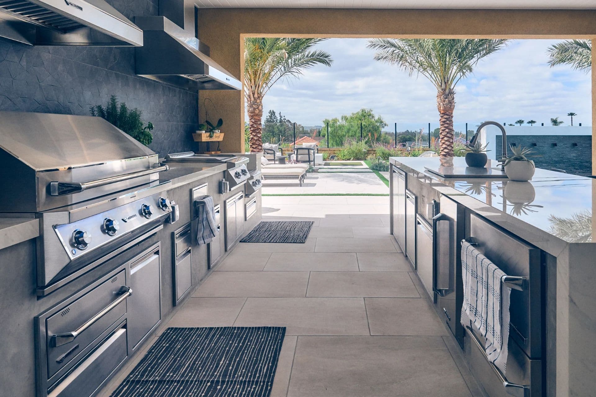 Sleek outdoor kitchen with stainless steel appliances, flanked by palm trees. Overlooks a pool and patio, creating a serene and modern ambiance.