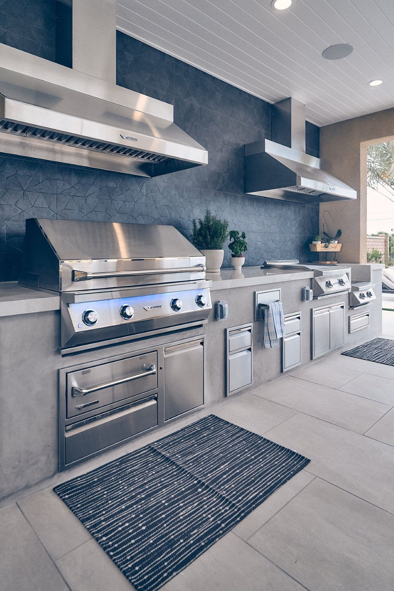 Modern outdoor kitchen with stainless steel appliances, including grills and storage. Potted plants decorate a sleek counter, creating a luxurious feel.