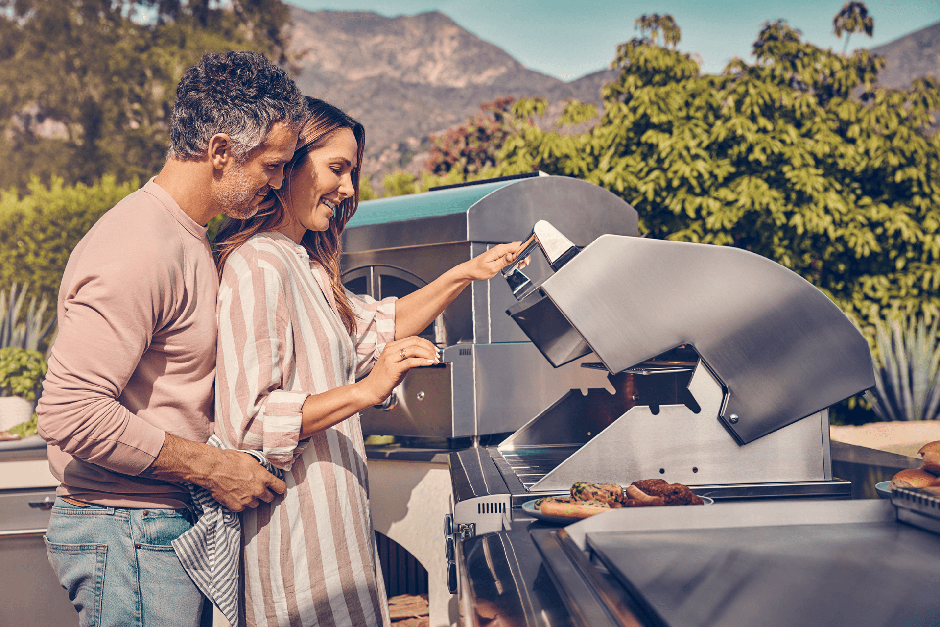A couple stands together at an outdoor grill, smiling as they cook. Surrounded by lush greenery and mountains, the scene is warm and inviting.