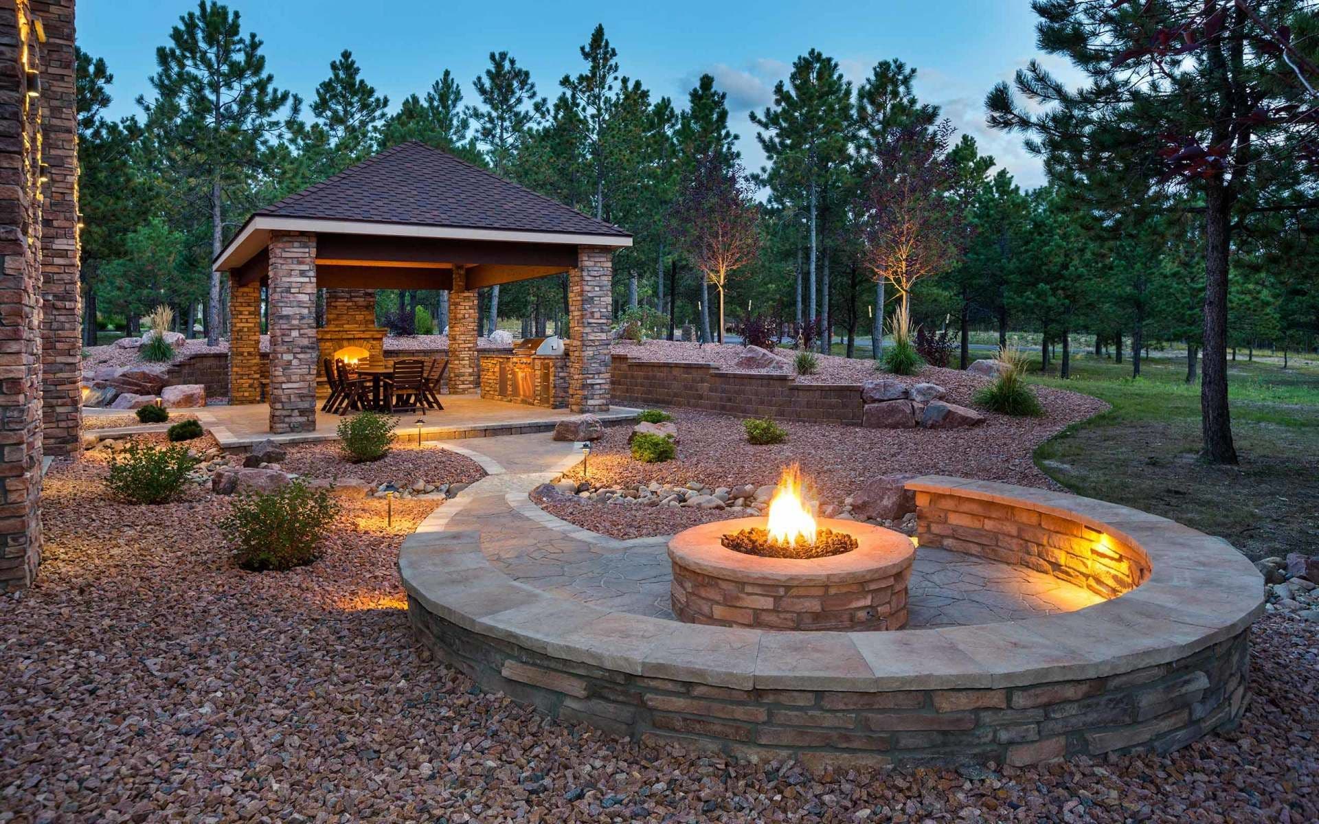 Cozy outdoor patio with a stone fire pit glowing warmly. A pavilion with a dining area is nestled among tall pine trees.