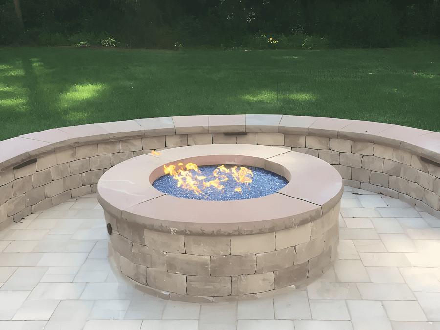 A circular stone fire pit with blue glass stones and orange flames, surrounded by a curved stone bench on a paved patio, with a lush green lawn beyond.