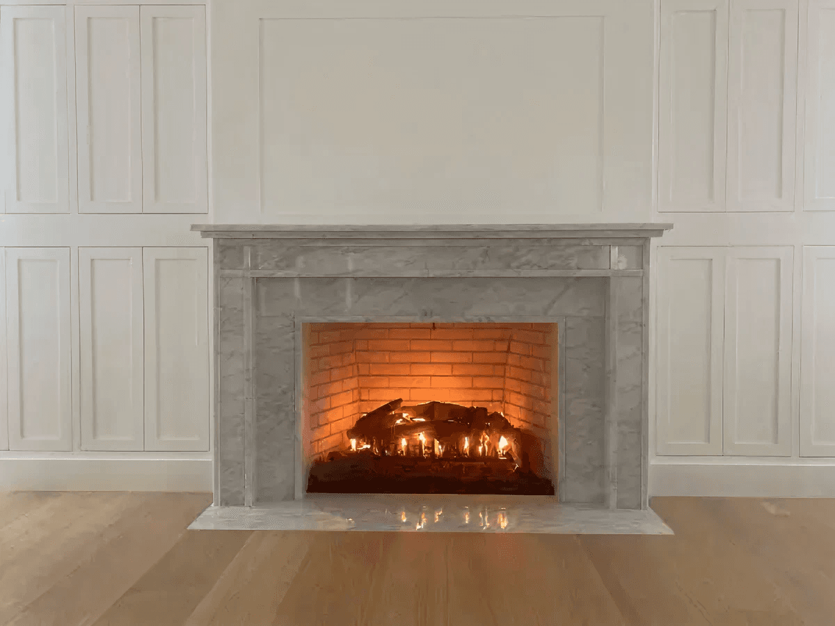 A cozy, lit fireplace with a marble mantel set against a white paneled wall. The warm glow contrasts with the polished wooden floor, creating an inviting ambiance.