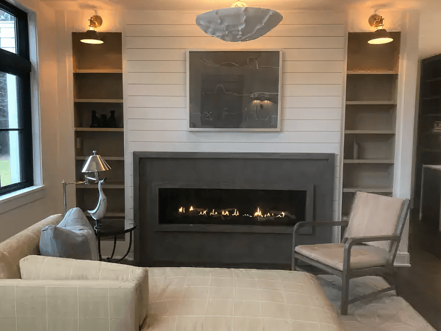 A modern living room with a lit fireplace centered on a white shiplap wall. Surrounding shelves, soft lighting, cozy seating, and neutral tones create a warm ambiance.