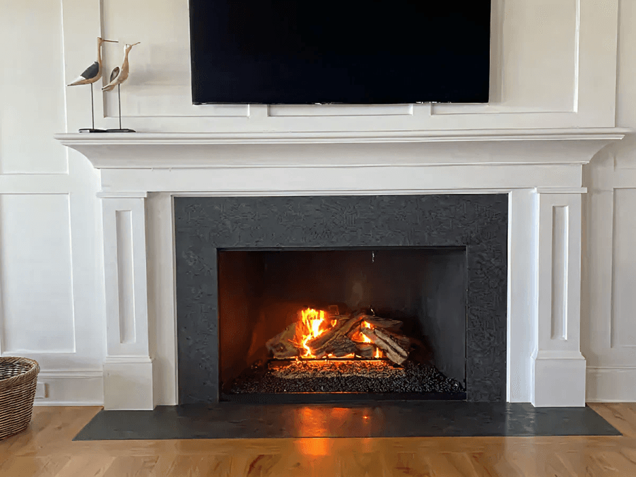 A cozy fireplace with burning logs under a white mantel. Above, a black TV contrasts the light walls. A basket and decorative birds add charm.