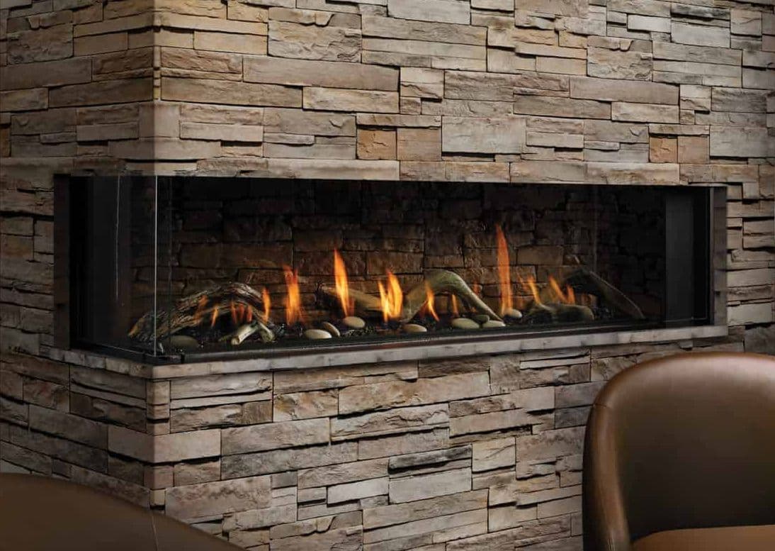 Modern stone fireplace with glass panel showcasing vivid orange flames and decorative logs. Cozy ambiance with a brown leather chair nearby.