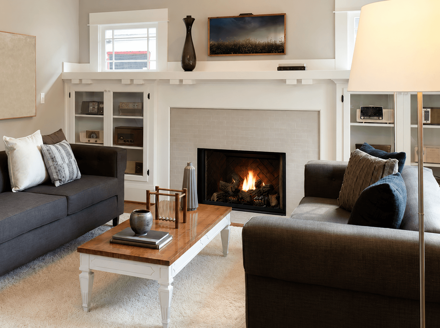A cozy living room with gray sofas, a wooden coffee table, and a lit fireplace. Shelves flank the fireplace, and a lamp casts a warm glow.