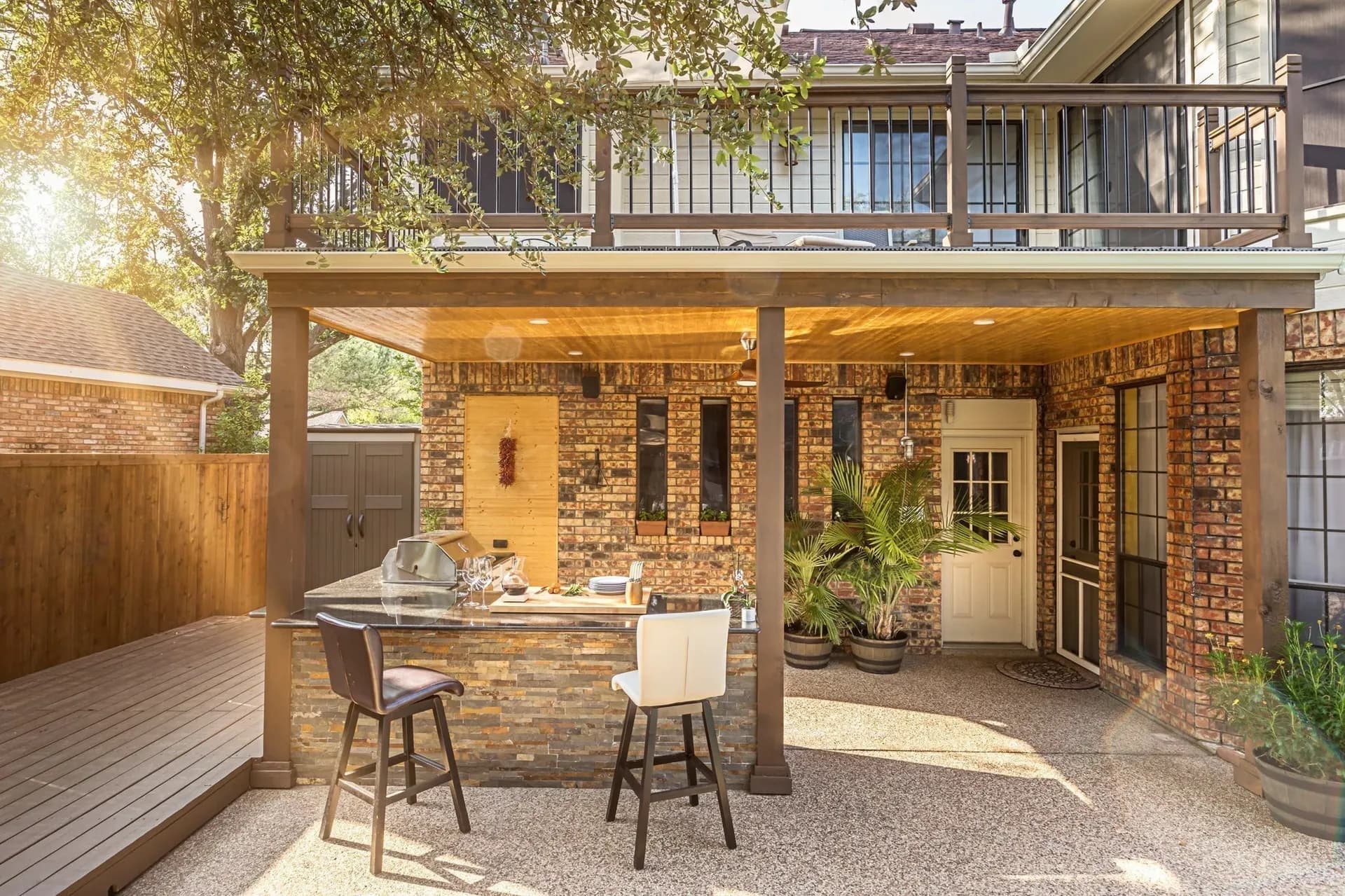 Outdoor patio with a stone bar, two stools, and potted plants under a wooden pergola. Sunlight filters through trees, creating a warm ambiance.