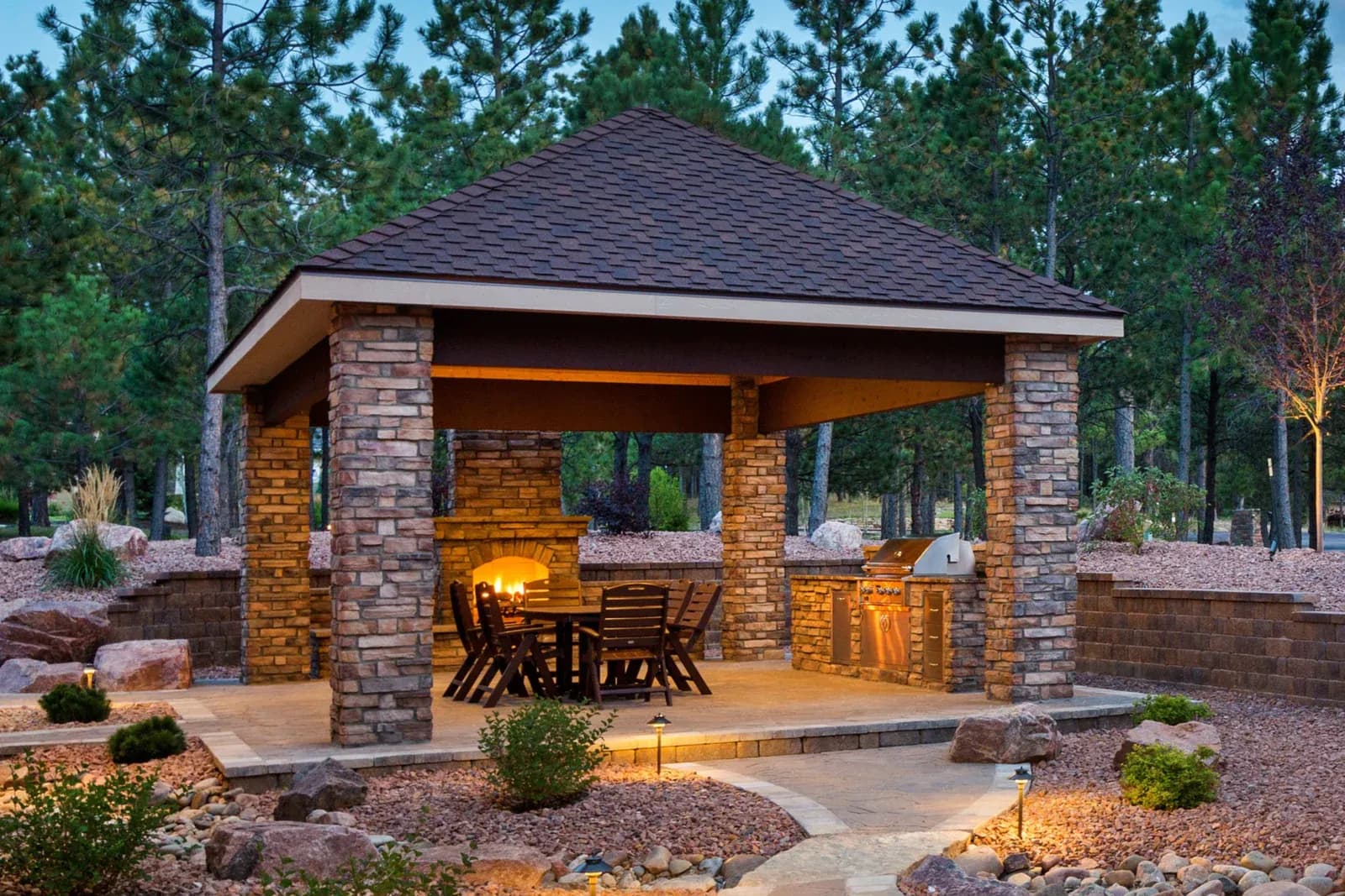 A cozy outdoor pavilion with a pitched roof and stone pillars. It features a warm-lit dining table and a barbecue grill, surrounded by trees and a landscaped patio.