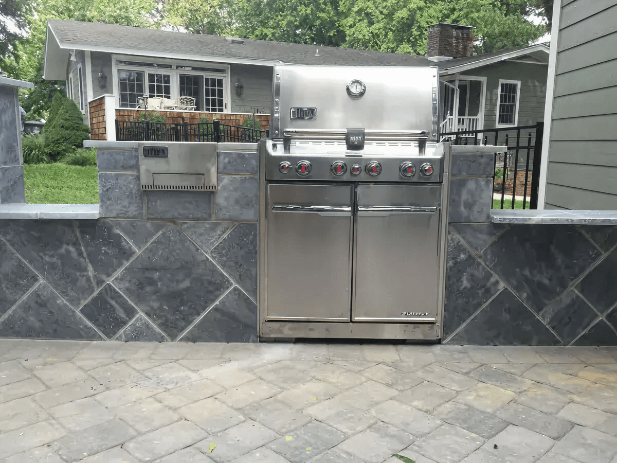 A stainless steel grill is set in a sleek, stone-tiled outdoor kitchen. The background shows a suburban home with a lush, green yard.