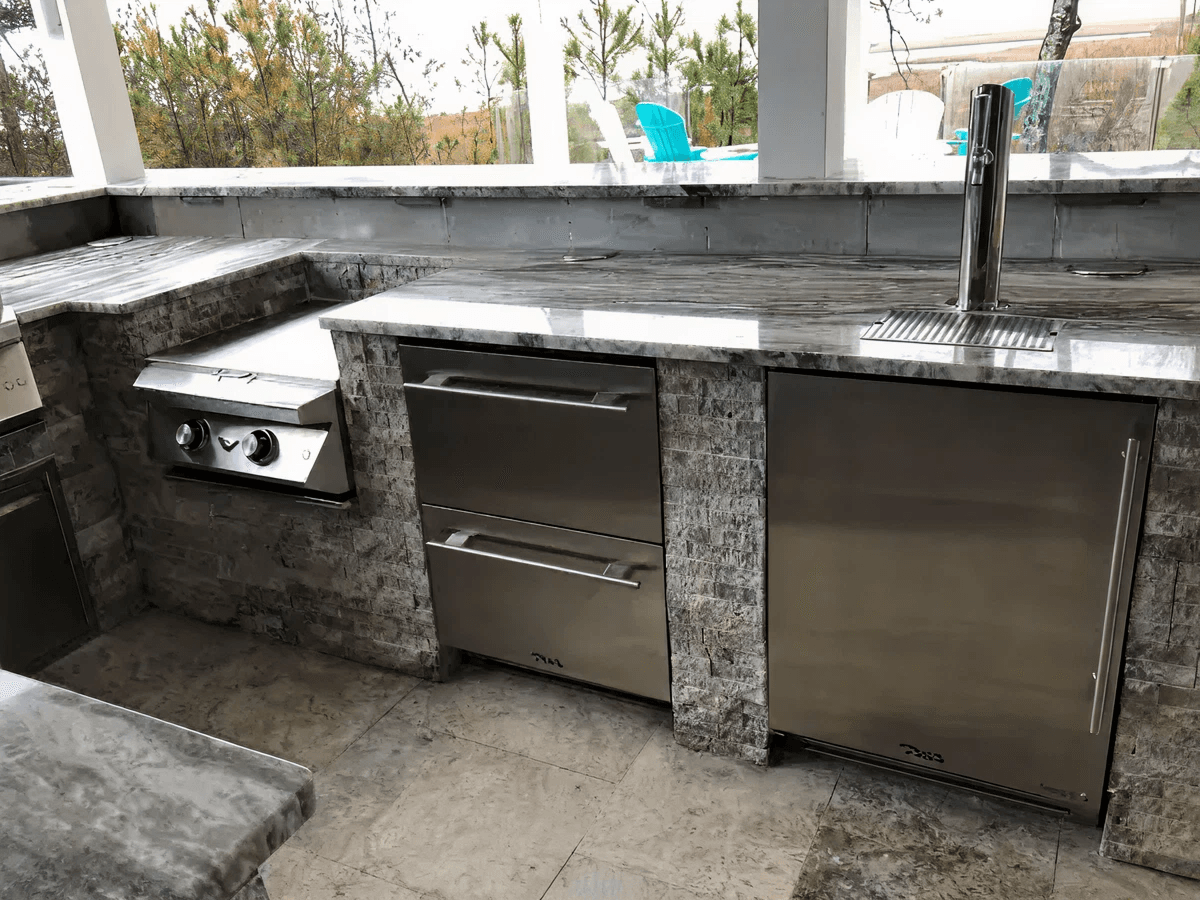 Outdoor kitchen with stainless steel appliances, a built-in grill, two drawers, and a fridge. Stone surfaces convey a sleek, modern design.