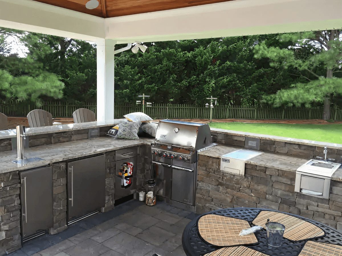 Outdoor kitchen with a stone countertop and built-in grill under a wooden pergola. Surrounded by trees, it conveys a cozy, inviting atmosphere.