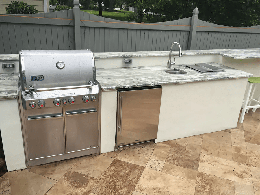 Outdoor kitchen setup with a stainless steel grill, marble countertop, sink, and mini fridge. The scene is bright with neutral tones and a relaxed ambiance.