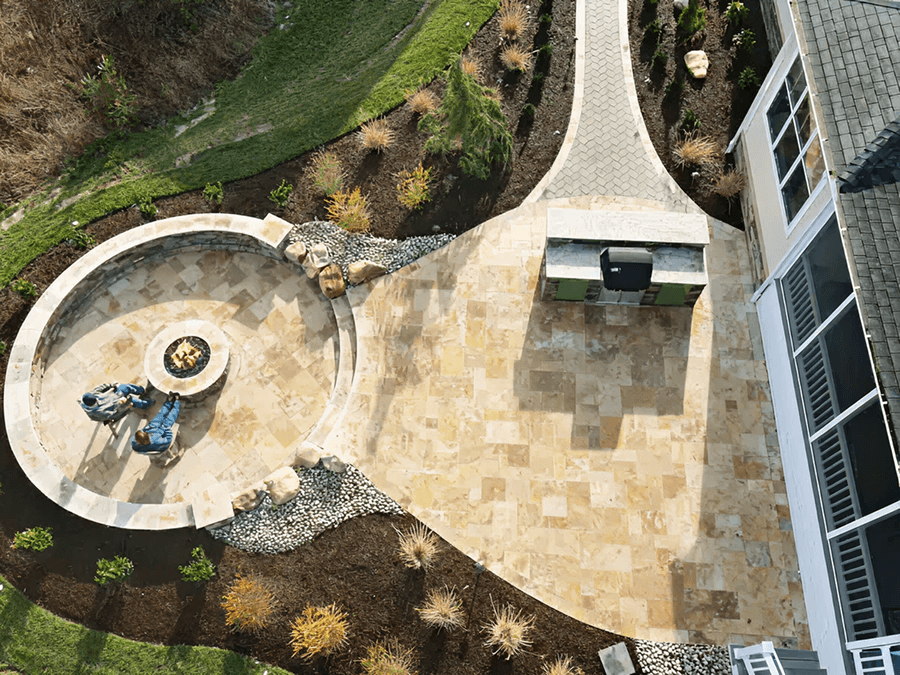 Aerial view of a landscaped backyard with a circular stone fire pit area featuring blue chairs, a stone path, barbecue grill, and neatly arranged shrubs.
