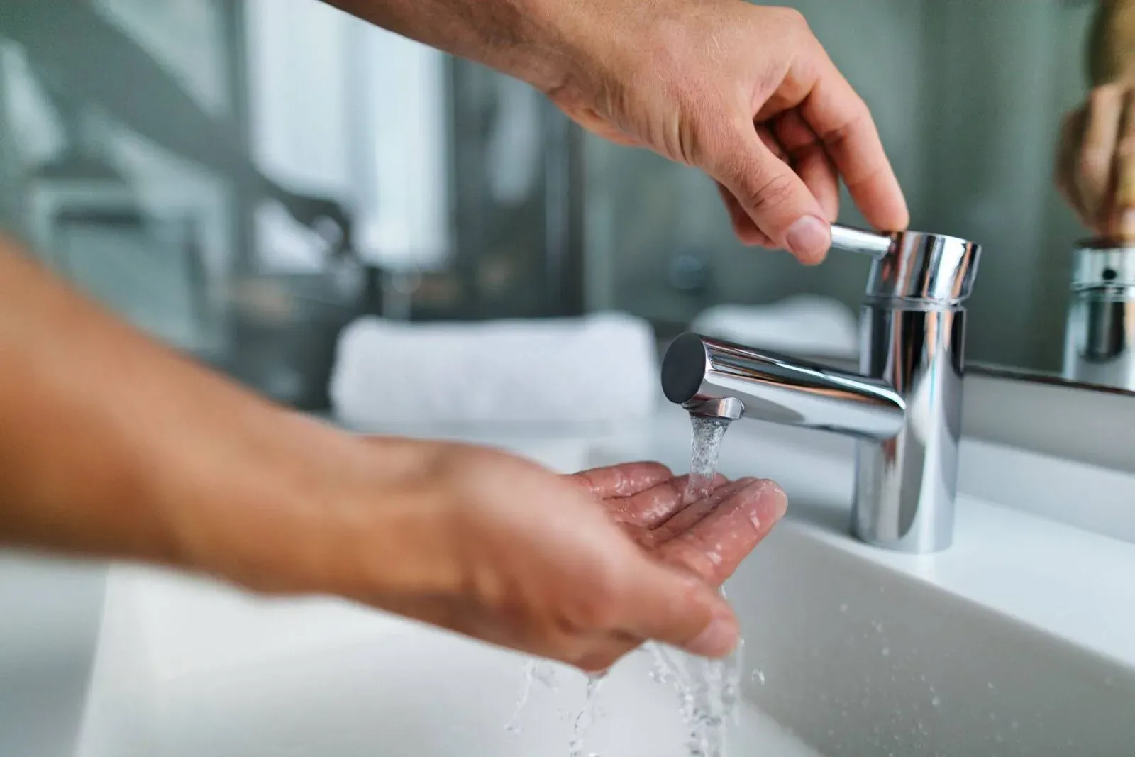 Hands adjusting a shiny chrome faucet over a white sink. Water flows gently, creating a clean, refreshing atmosphere in a modern bathroom setting.