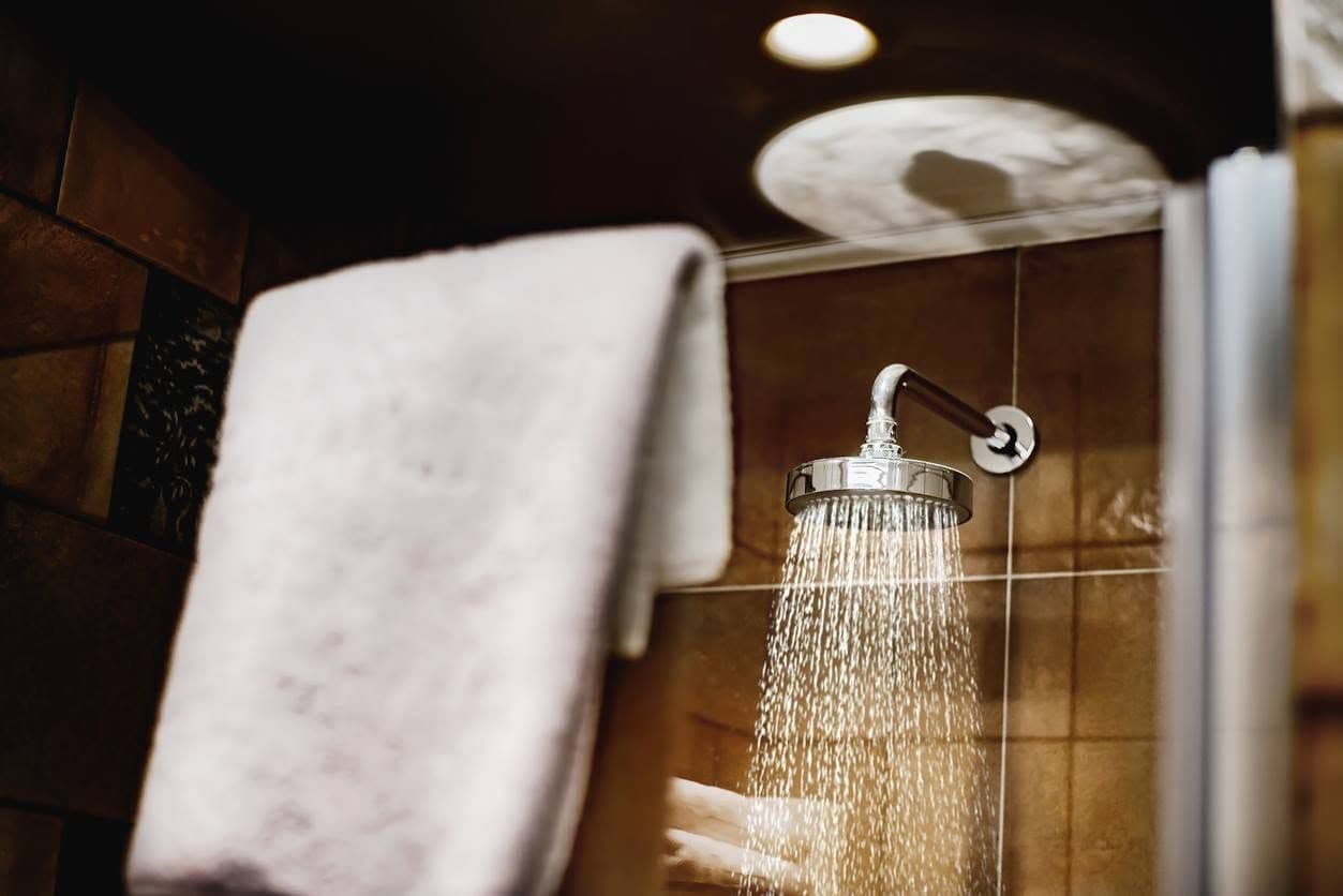 Showerhead with flowing water in a bathroom, casting a shadow. A towel hangs on a rail to the left.