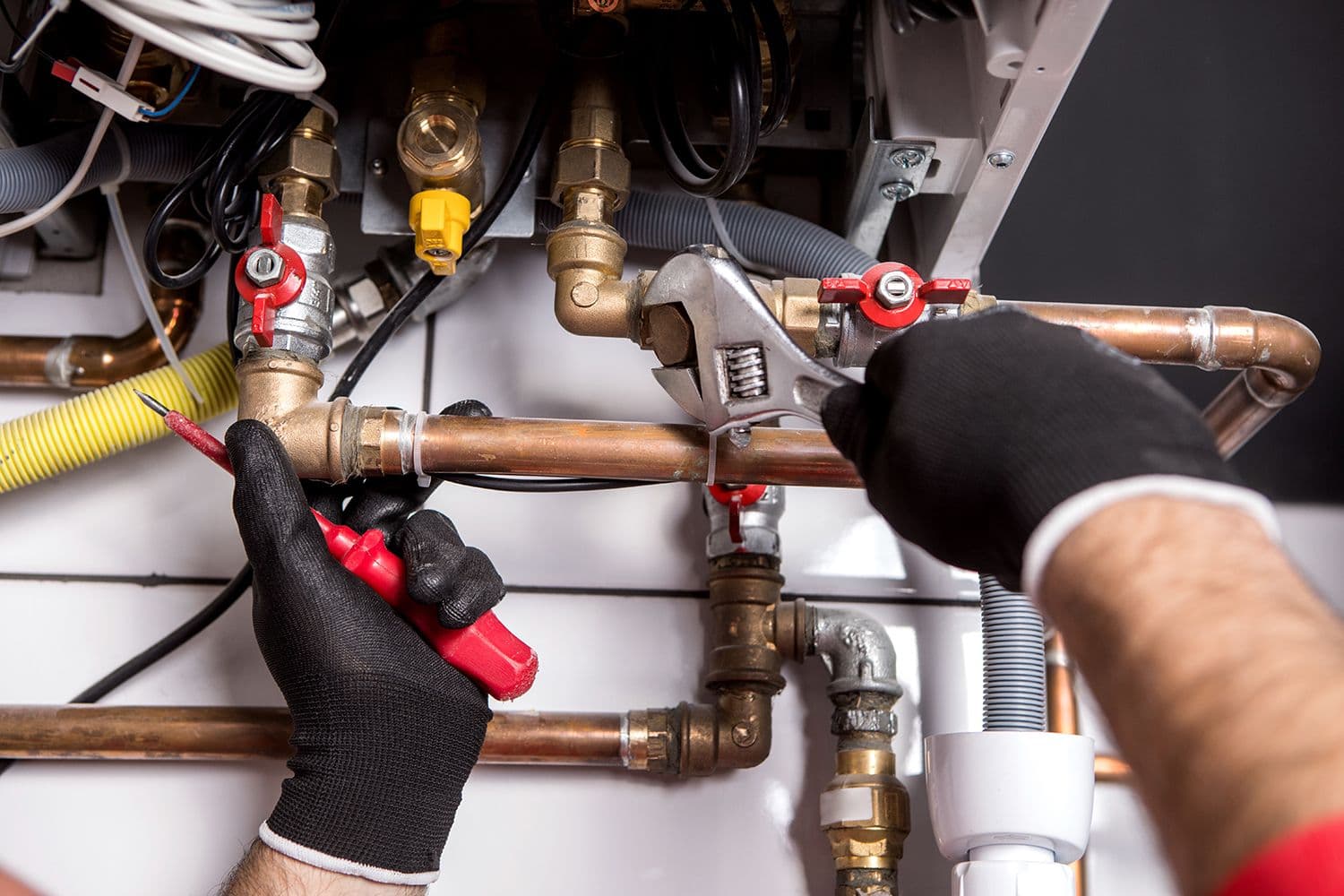 A person wearing gloves adjusts copper pipes using a wrench and screwdriver. The setting includes various valves and fittings in a mechanical room.