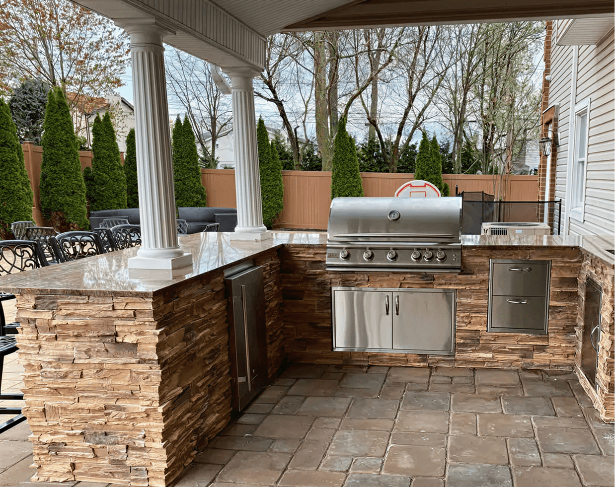 Outdoor kitchen with a stone countertop, stainless steel grill, and drawers. Surrounded by trees, a privacy fence, and patio seating, creating a cozy, elegant setting.